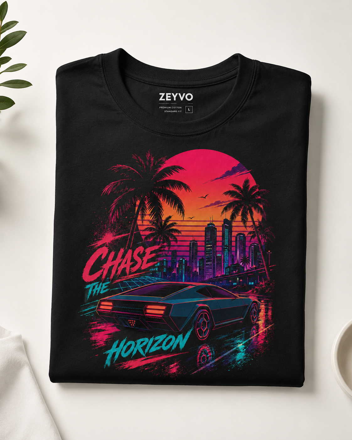 A folded black t-shirt with a colorful retro-futuristic graphic print featuring a car, palm trees, a city skyline, and neon text, displayed on a light surface with a plant leaf and a cup edge visible.