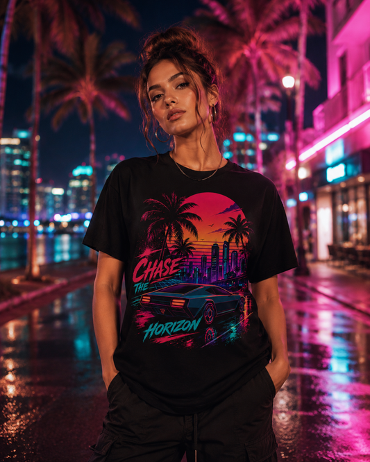 A woman wearing a black graphic t-shirt with a retro 80s synthwave design featuring a car, palm trees, and neon text, standing on a wet city street at night with palm trees and neon lights in the background.