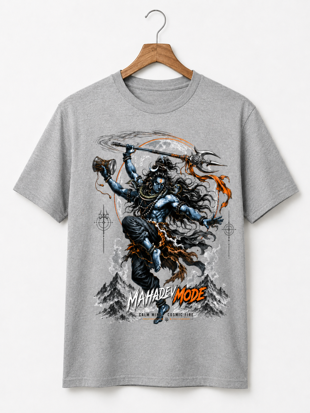 A heather gray short-sleeve t-shirt hanging on a wooden hanger against a light gray background. The shirt features a large graphic print of a Hindu deity with a trident and mace, set against a circular moon and mountain background, with the text 'MAHADEI MODE' in orange and white.
